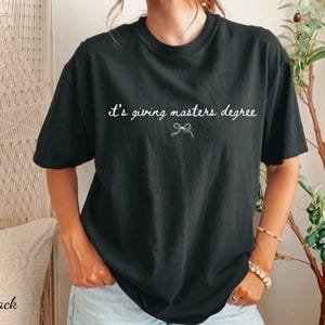 Puede incluir: Camiseta negra con la frase "it's giving masters degree" en escritura blanca. Un peque&ntilde;o lazo gr&aacute;fico est&aacute; debajo del texto. La camiseta es de cuello redondo y tiene mangas cortas.