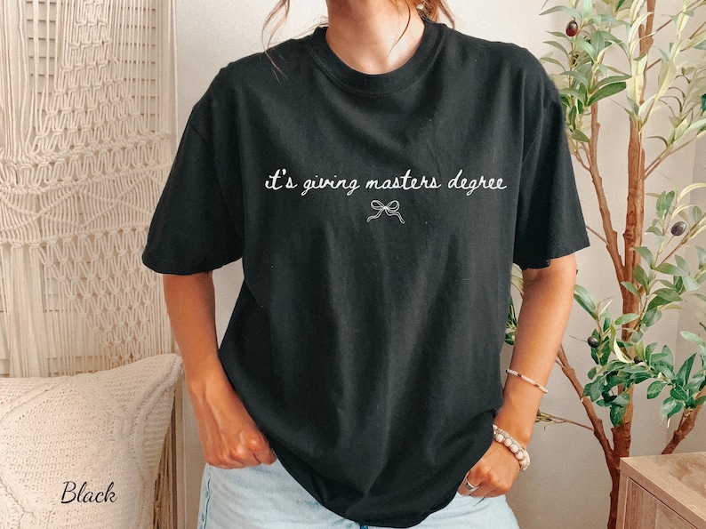 Puede incluir: Camiseta negra con la frase "it's giving masters degree" en escritura blanca. Un peque&ntilde;o lazo gr&aacute;fico est&aacute; debajo del texto. La camiseta es de cuello redondo y tiene mangas cortas.