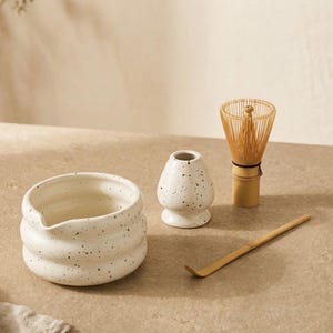May include: A white ceramic matcha bowl with a speckled pattern and a pouring spout, a matching white speckled ceramic whisk holder, a light brown bamboo matcha whisk, and a long, thin, curved bamboo matcha scoop for preparing tea.