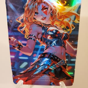 May include: A holographic trading card featuring a stylized illustration of a woman with long, flowing blonde hair and blue eyes. She wears a black and silver outfit with red accents. The card is titled "Pleiades, Superstar, Revered Guardian Hero."