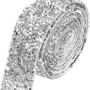 May include: A roll of silver-toned adhesive trim featuring a dense, multi-faceted surface of clear, sparkling rhinestones. The flexible strip is coiled into a circular shape, showcasing a continuous, reflective texture across the entire length.