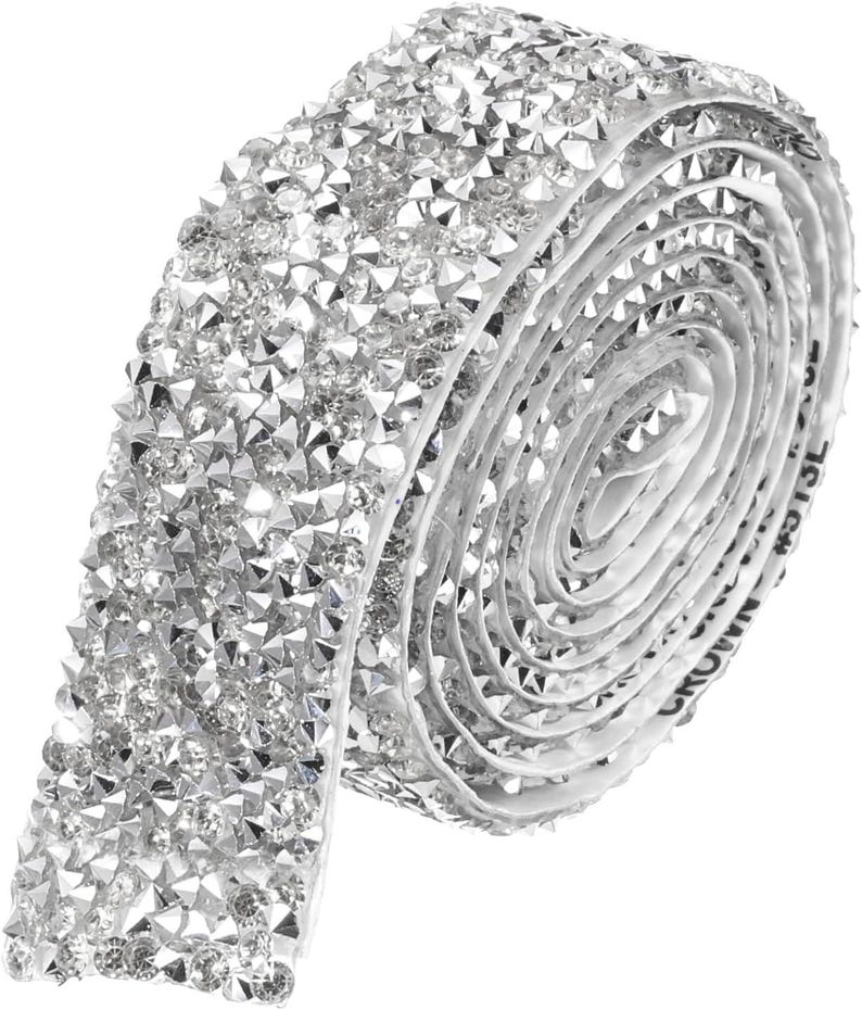 May include: A roll of silver-toned adhesive trim featuring a dense, multi-faceted surface of clear, sparkling rhinestones. The flexible strip is coiled into a circular shape, showcasing a continuous, reflective texture across the entire length.