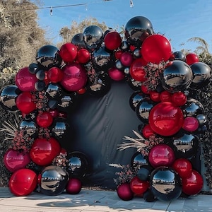 May include: A balloon arch featuring a mix of red, black, and silver balloons. The balloons are arranged in an arch shape, with some floral accents. The backdrop is a dark fabric, and the scene is outdoors.