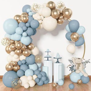 May include: A decorative balloon arch in shades of blue, gold, and cream, with a teddy bear and dessert stands. The balloons are arranged in an arch shape, with some clear balloons filled with gold confetti.