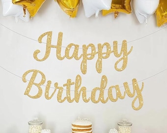 May include: A festive birthday scene with gold glitter "Happy Birthday" banner, gold and white balloons, a cake, and snacks. The banner is strung across a white wall, with a cake and treats on a table below. The balloons add a celebratory touch.