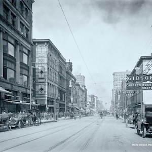 Könnte beinhalten: Ein Schwarzweißfoto einer Straßenszene in Memphis, Tennessee. Das Foto zeigt eine Straße, die von Gebäuden gesäumt ist, mit einem Schild für "Gibson's Carpets Rugs Draperies" auf der rechten Seite des Bildes. Das Foto wurde 1907 aufgenommen.