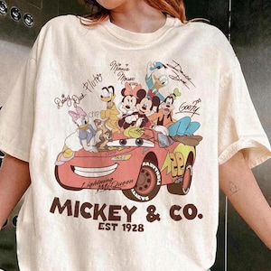 Może przedstawiać: Kremowa koszulka z grafiką postaci Disneya, w tym Myszki Miki, Myszki Minnie, Kaczora Donalda i Goofy'ego. Projekt zawiera czerwony samoch&oacute;d z napisem "Lightning McQueen" i "Mickey & Co. EST 1928."