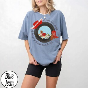 May include: A light blue t-shirt featuring a graphic of a black tire framing a sky blue background with a red cowboy hat, a red cowboy boot, and cartoon characters. The text reads, Love has ways of bringing things back to life, with Taylor written on the boot.