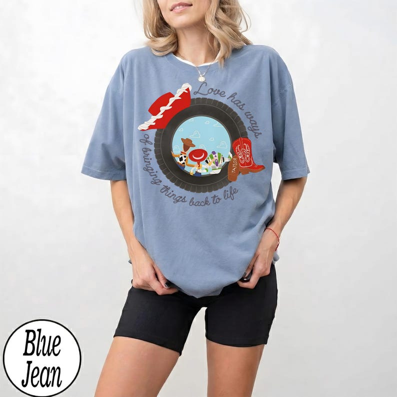 May include: A light blue t-shirt featuring a graphic of a black tire framing a sky blue background with a red cowboy hat, a red cowboy boot, and cartoon characters. The text reads, Love has ways of bringing things back to life, with Taylor written on the boot.