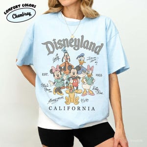 K&ouml;nnte beinhalten: Hellblaues T-Shirt mit einem Disneyland-Motiv mit Zeichentrickfiguren wie Mickey Mouse, Minnie Mouse und Goofy. Das Shirt enth&auml;lt den Text "Disneyland California" und "EST. 1955". Das Shirt ist aus Chambray Comfort Colors.