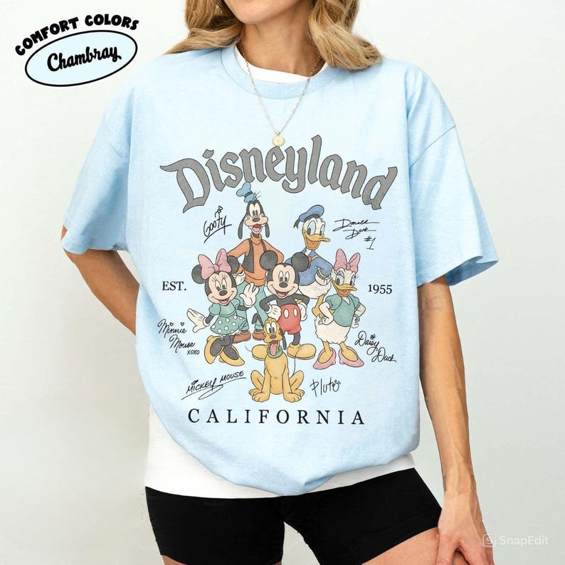 K&ouml;nnte beinhalten: Hellblaues T-Shirt mit einem Disneyland-Motiv mit Zeichentrickfiguren wie Mickey Mouse, Minnie Mouse und Goofy. Das Shirt enth&auml;lt den Text "Disneyland California" und "EST. 1955". Das Shirt ist aus Chambray Comfort Colors.