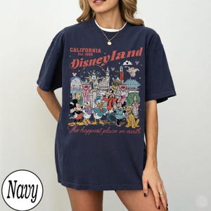 May include: Navy blue t-shirt with a Disneyland graphic. The design includes the text "CALIFORNIA Est. 1955 Disneyland" and "The happiest place on earth" with cartoon characters and park landmarks.