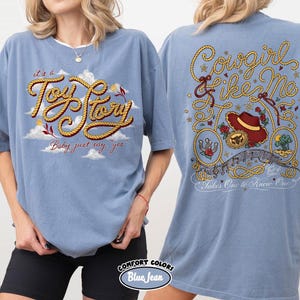 May include: A blue denim-colored t-shirt featuring a front graphic with the text 'it's a Toy Story Baby just say yes' in rope-style lettering amidst clouds. The back graphic displays 'Cowgirl like Me' and 'Takes One to Know One' with rope accents, a cowboy hat, and floral motifs.