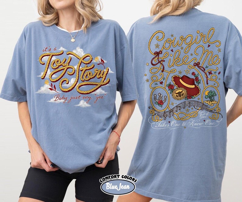 May include: A blue denim-colored t-shirt featuring a front graphic with the text 'it's a Toy Story Baby just say yes' in rope-style lettering amidst clouds. The back graphic displays 'Cowgirl like Me' and 'Takes One to Know One' with rope accents, a cowboy hat, and floral motifs.