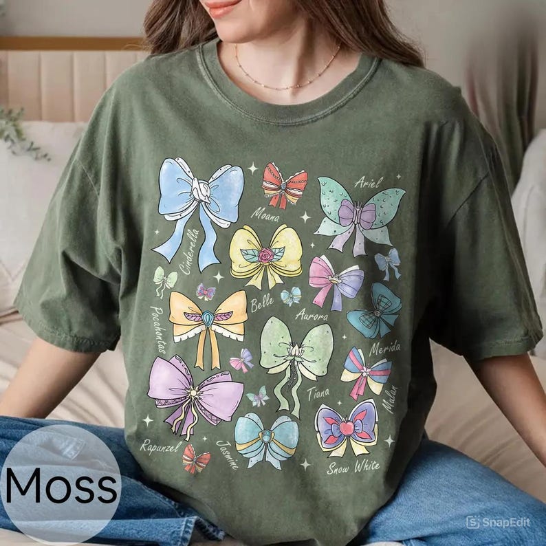 Peut inclure: T-shirt vert mousse avec des illustrations d'arcs color&eacute;s, chacun portant le nom d'une princesse Disney. Les n&oelig;uds sont de diff&eacute;rentes couleurs, dont le bleu, le rose, le jaune et le violet. Le t-shirt a une coupe d&eacute;contract&eacute;e.