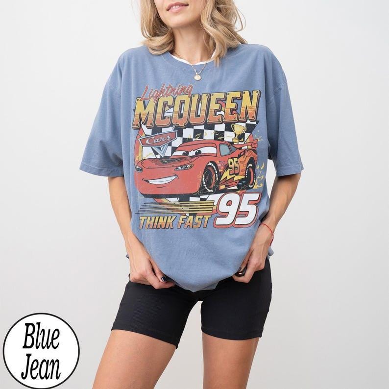 May include: A light blue t-shirt featuring a graphic of Lightning McQueen from the Cars movie. The design includes the text "Lightning McQueen", "Cars", "95", and "Think Fast". The shirt is paired with black shorts.