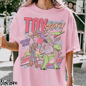 May include: Pink t-shirt featuring the words "Toy Story" in a retro font, with Woody, Buzz Lightyear, and Rex the dinosaur. The shirt also includes the text "Since 1995" and the brand name "Blossom".