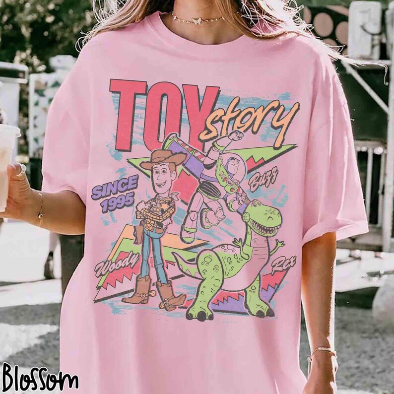 May include: Pink t-shirt featuring the words "Toy Story" in a retro font, with Woody, Buzz Lightyear, and Rex the dinosaur. The shirt also includes the text "Since 1995" and the brand name "Blossom".