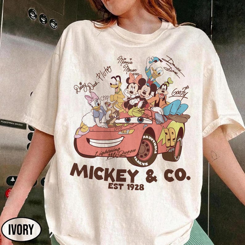 Może przedstawiać: Kremowa koszulka z grafiką postaci Disneya, w tym Myszki Miki, Myszki Minnie, Kaczora Donalda i Goofy'ego. Projekt zawiera czerwony samoch&oacute;d z napisem "Lightning McQueen" i "Mickey & Co. EST 1928."