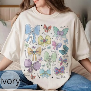 Peut inclure: T-shirt ivoire avec un motif de n&oelig;uds color&eacute;s, chacun portant le nom d'une princesse Disney. Les n&oelig;uds sont dans des tons pastel de bleu, rose et jaune. Le t-shirt a une coupe d&eacute;contract&eacute;e.