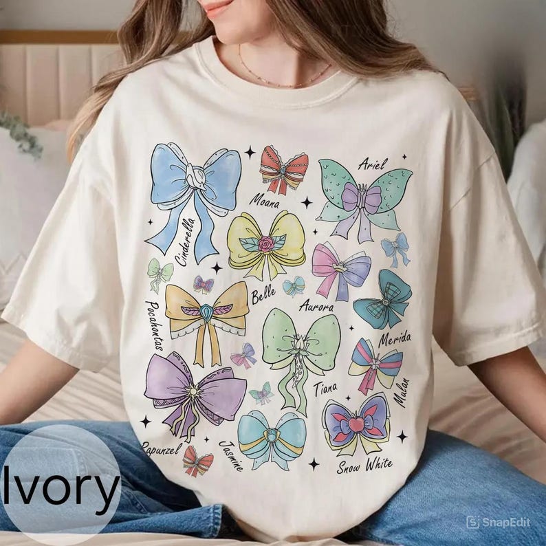 Peut inclure: T-shirt ivoire avec un motif de n&oelig;uds color&eacute;s, chacun portant le nom d'une princesse Disney. Les n&oelig;uds sont dans des tons pastel de bleu, rose et jaune. Le t-shirt a une coupe d&eacute;contract&eacute;e.
