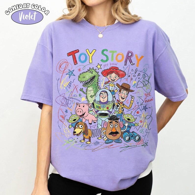 May include: A violet short-sleeve t-shirt featuring a colorful graphic of Toy Story characters including Woody, Buzz Lightyear, Jessie, Rex, Hamm, Mr. Potato Head, Slinky Dog, and Aliens, surrounded by multi-colored scribbles and the text Toy Story.