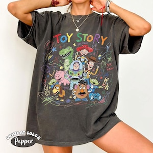 May include: A charcoal gray short-sleeve t-shirt featuring a colorful graphic print of Toy Story characters including Woody, Buzz Lightyear, Jessie, Rex, Hamm, Slinky Dog, Mr. Potato Head, and Aliens, surrounded by multicolored scribbled crayon-like lines.