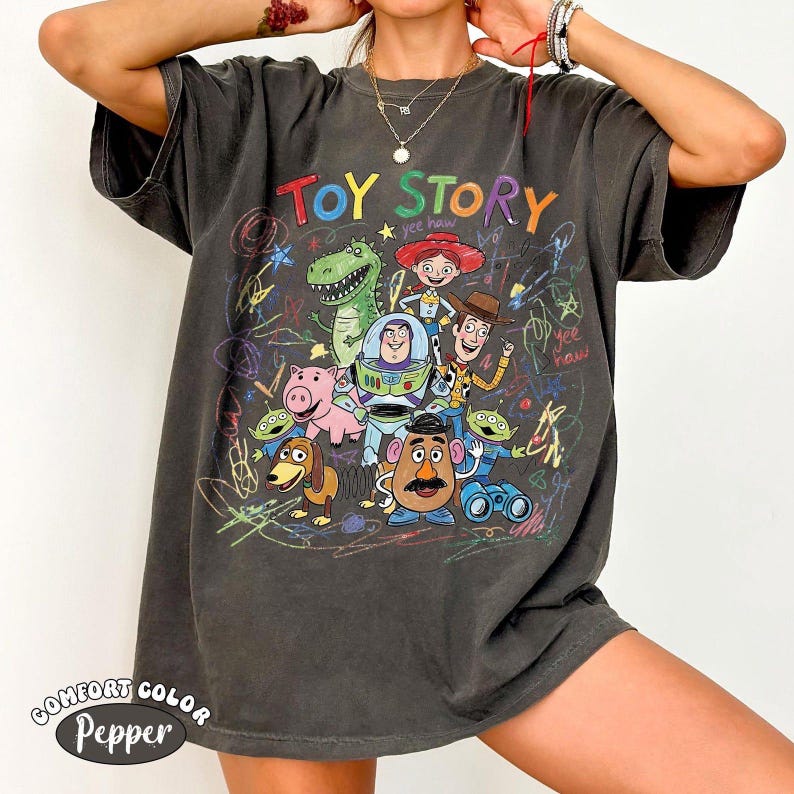 May include: A charcoal gray short-sleeve t-shirt featuring a colorful graphic print of Toy Story characters including Woody, Buzz Lightyear, Jessie, Rex, Hamm, Slinky Dog, Mr. Potato Head, and Aliens, surrounded by multicolored scribbled crayon-like lines.