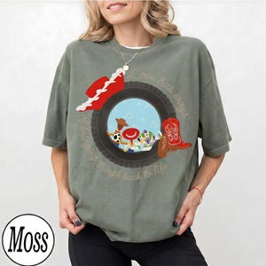 May include: A moss green short-sleeve t-shirt featuring a graphic of a black tire framing cartoon characters. The design includes a red cowboy hat, a red cowboy boot, and the text "Love has ways of bringing things back to life" in script.
