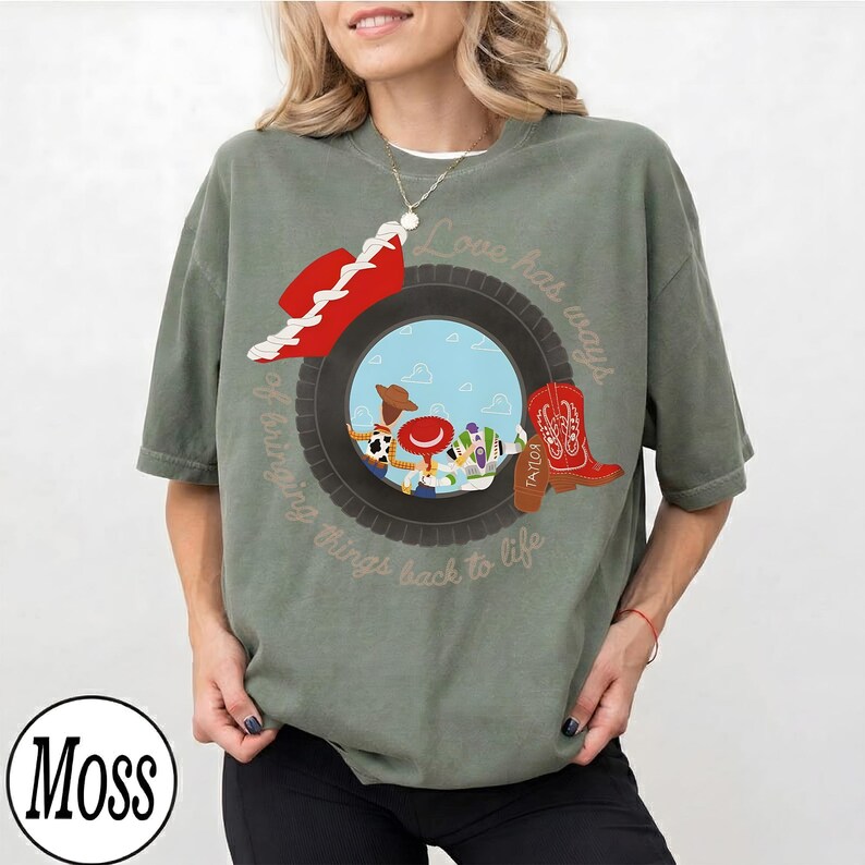 May include: A moss green short-sleeve t-shirt featuring a graphic of a black tire framing cartoon characters. The design includes a red cowboy hat, a red cowboy boot, and the text "Love has ways of bringing things back to life" in script.