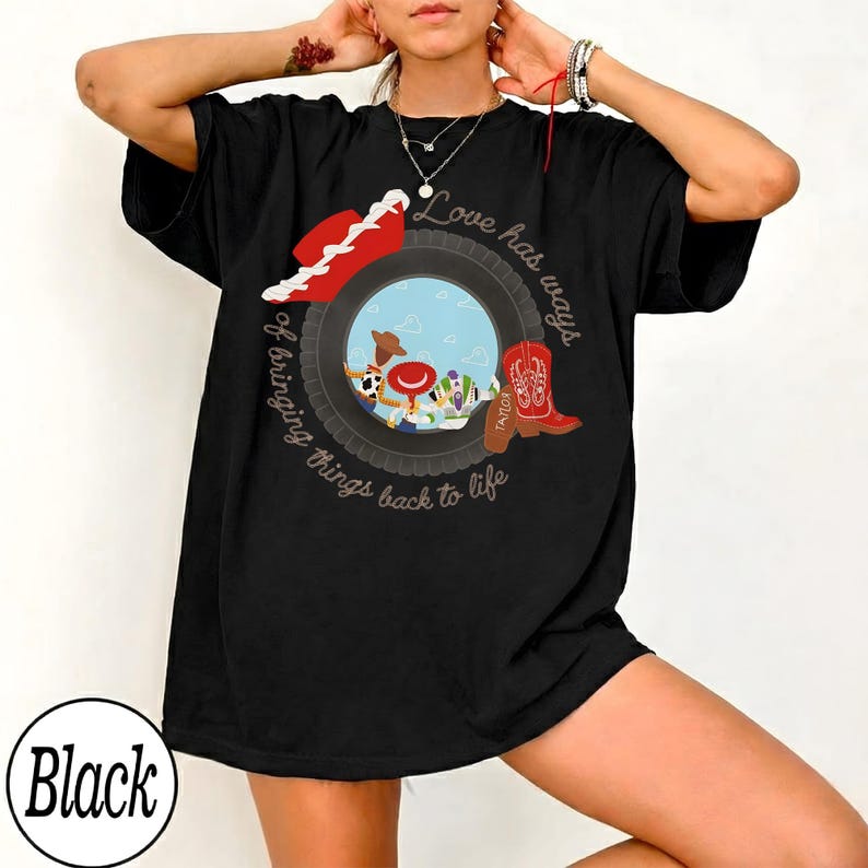 May include: A black short-sleeve t-shirt featuring a graphic of a tire framing cartoon characters, a red cowboy hat, and a red cowboy boot. The text reads, "Love has ways of bringing things back to life" in a circular script around the central tire design.