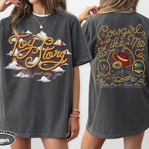 May include: A charcoal gray t-shirt featuring a front graphic with the text 'it's a Toy Story' in rope-style lettering amidst white clouds. The back graphic displays 'Cowgirl like me' in rope text, a red hat, a horseshoe, and a desert scene.
