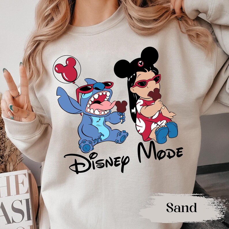 May include: Sand-colored sweatshirt featuring a graphic of Stitch and Lilo from Disney's Lilo & Stitch, both wearing sunglasses and holding ice cream. The words "Disney Mode" are printed below the characters.