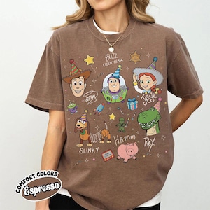 May include: An espresso brown short-sleeve t-shirt featuring a graphic print of Toy Story characters including Woody, Buzz Lightyear, Jessie, Slinky, Hamm, and Rex wearing party hats. The design includes stars, a rocket, and handwritten names.
