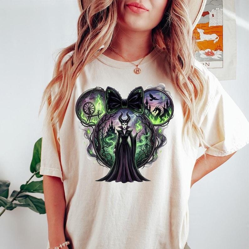May include: Cream-colored t-shirt featuring a graphic of a villainous character in a black dress, surrounded by green flames and a bow. The design incorporates elements of a fairytale theme, with a castle and dragon. The shirt has a round neckline.