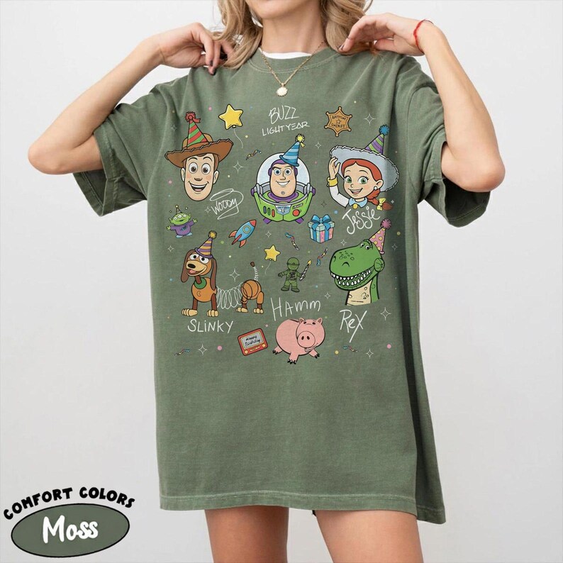 May include: An olive green t-shirt featuring a graphic print of Toy Story characters including Woody, Buzz Lightyear, Jessie, Slinky Dog, Rex, and Hamm wearing party hats. The design includes text labels for each character and small festive icons.