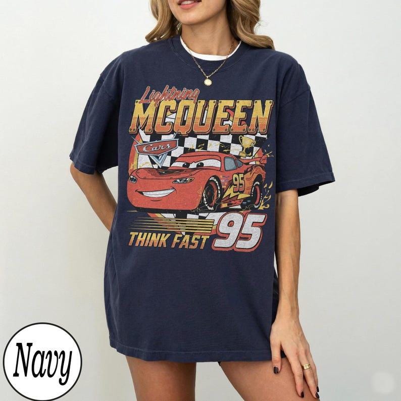 May include: Navy blue t-shirt featuring a graphic of Lightning McQueen from the Cars movie. The design includes the text "Lightning McQueen," "Cars," and "Think Fast 95." The graphic is in red, yellow, and white.