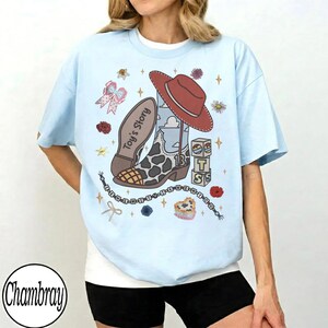 Puede incluir: Una camiseta azul claro con un estampado gr&aacute;fico de una bota de vaquero con patr&oacute;n de vaca, un sombrero de vaquero rojo, bloques con las letras T, S y una estrella, y el texto Toy's Story. El dise&ntilde;o incluye peque&ntilde;as flores, estrellas y cuentas.