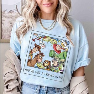 Puede incluir: Camiseta azul claro con un gr&aacute;fico colorido de personajes de la pel&iacute;cula animada "Toy Story". El gr&aacute;fico incluye a Woody, Buzz Lightyear y otros personajes. El texto "YOU'VE GOT A FRIEND IN ME" est&aacute; impreso debajo.