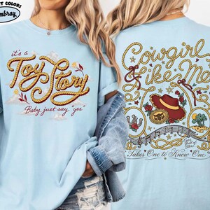 May include: A light blue t-shirt featuring a front graphic with yellow rope lettering that reads "it's a Toy Story Baby, just say "yes"". The back graphic displays "Cowgirl like Me" in rope text, a cowboy hat, a belt buckle, and musical notes.