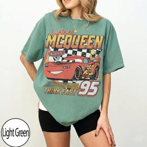 May include: Light green t-shirt featuring Lightning McQueen from the Cars movie. The graphic includes the text "Lightning McQueen", "Cars", "Think Fast", and the number "95". The design is in shades of red, orange, yellow, and black.