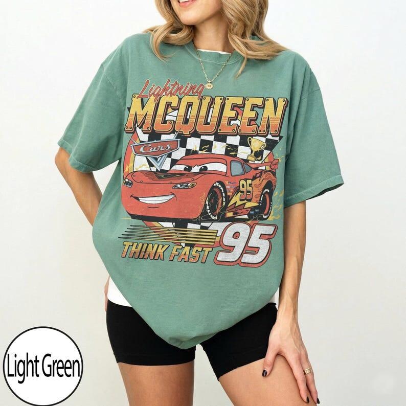 May include: Light green t-shirt featuring Lightning McQueen from the Cars movie. The graphic includes the text "Lightning McQueen", "Cars", "Think Fast", and the number "95". The design is in shades of red, orange, yellow, and black.