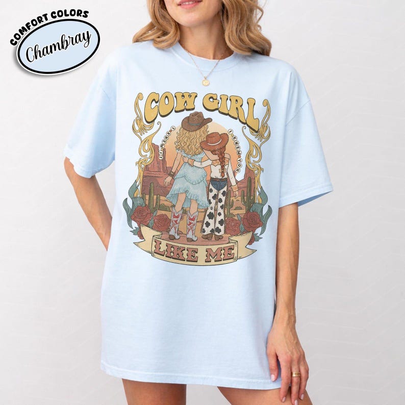 Puede incluir: Una camiseta azul claro con un estampado gr&aacute;fico de dos figuras con sombreros de vaquero en un paisaje des&eacute;rtico con cactus y rosas. El texto dice COW GIRL LIKE ME. La camiseta tiene cuello redondo y mangas cortas con etiqueta Comfort Colors.