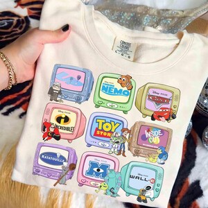 May include: Cream-colored sweatshirt featuring cartoon televisions with Disney Pixar movie graphics. The designs include "Finding Nemo," "Cars," "The Incredibles," "Toy Story," "Inside Out," "Ratatouille," "Monsters, Inc.," and "Wall-E."