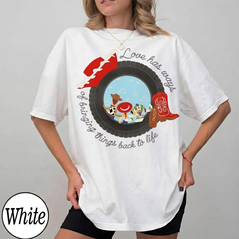 May include: A white short-sleeve t-shirt featuring a graphic of a black tire framing a sky-blue background with cartoon characters. The text "Love has ways of bringing things back to life" is printed in a circular script around the tire graphic.