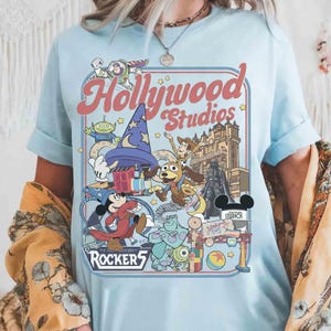K&ouml;nnte beinhalten: Hellblaues T-Shirt mit einer farbenfrohen Grafik von Disney's Hollywood Studios. Das Design zeigt Zeichentrickfiguren wie Mickey Mouse, Buzz Lightyear und Woody, mit den Worten "Hollywood Studios" und "Rockers" im Retro-Stil.
