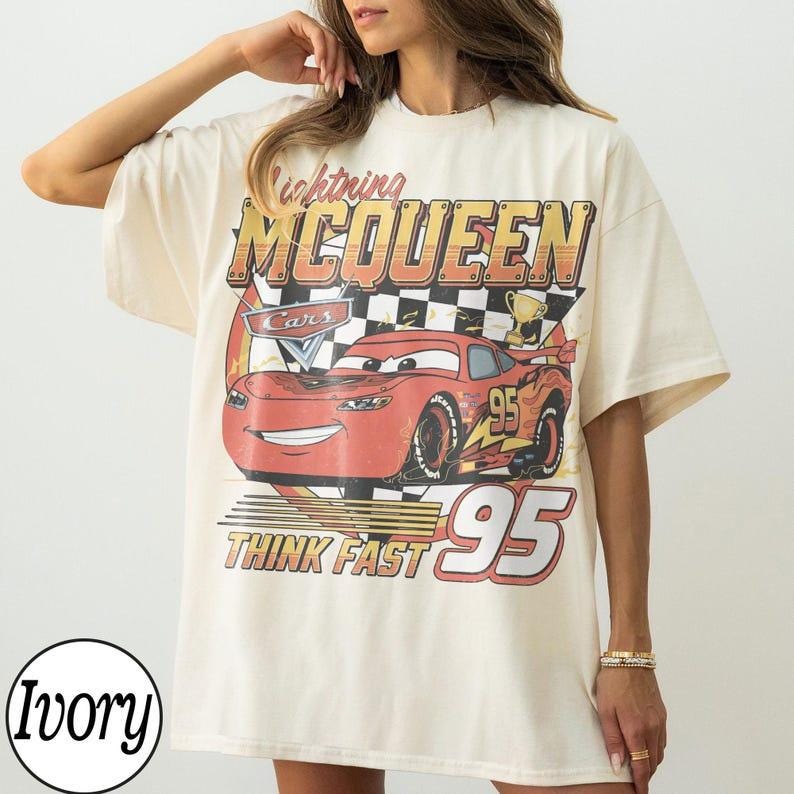 May include: Ivory-colored t-shirt featuring a graphic of Lightning McQueen from the movie Cars. The design includes the text "Lightning McQueen," "Cars," "95," and "Think Fast." The shirt has short sleeves and a relaxed fit.