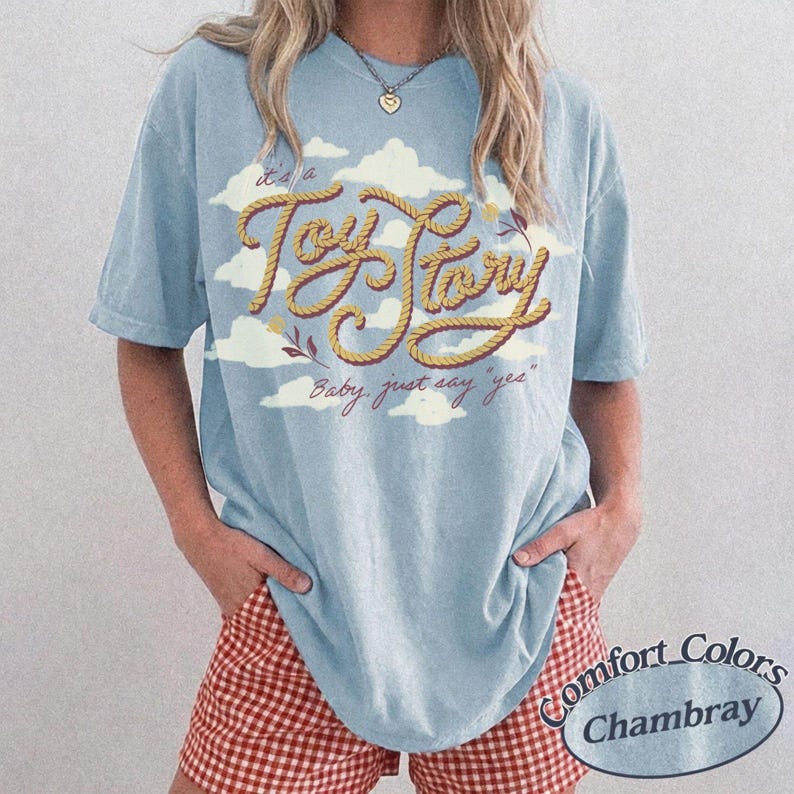 May include: A light blue short-sleeve t-shirt featuring a graphic of white clouds and the text "Toy Story" written in a yellow rope-style font. Below the main text, the phrase "Baby, just say 'yes'" is printed in a cursive red script font.