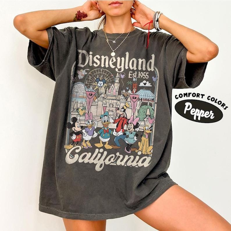 May include: Dark gray t-shirt featuring a Disneyland graphic with Mickey Mouse, Donald Duck, and other characters. The design includes the words "Disneyland California Est 1955" in a vintage style. The shirt is labeled "Comfort Colors Pepper".