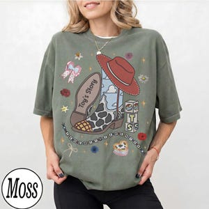 Puede incluir: Una camiseta verde musgo con un estampado gr&aacute;fico de una bota de vaquero con manchas de vaca, un sombrero de vaquero rojo, un lazo rosa y bloques apilados con las letras T, S y el texto Toy's Story, adem&aacute;s de estrellas y flores.